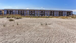 Un hotel de varios pisos con techo plano y ventanas amplias, ubicado en una playa de arena con poca vegetación.