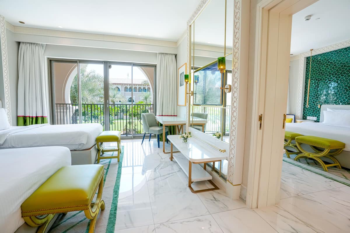 Une chambre d’hôtel avec deux lits, une table à manger, un banc, un grand miroir et un balcon donnant sur un espace verdoyant et un immeuble.