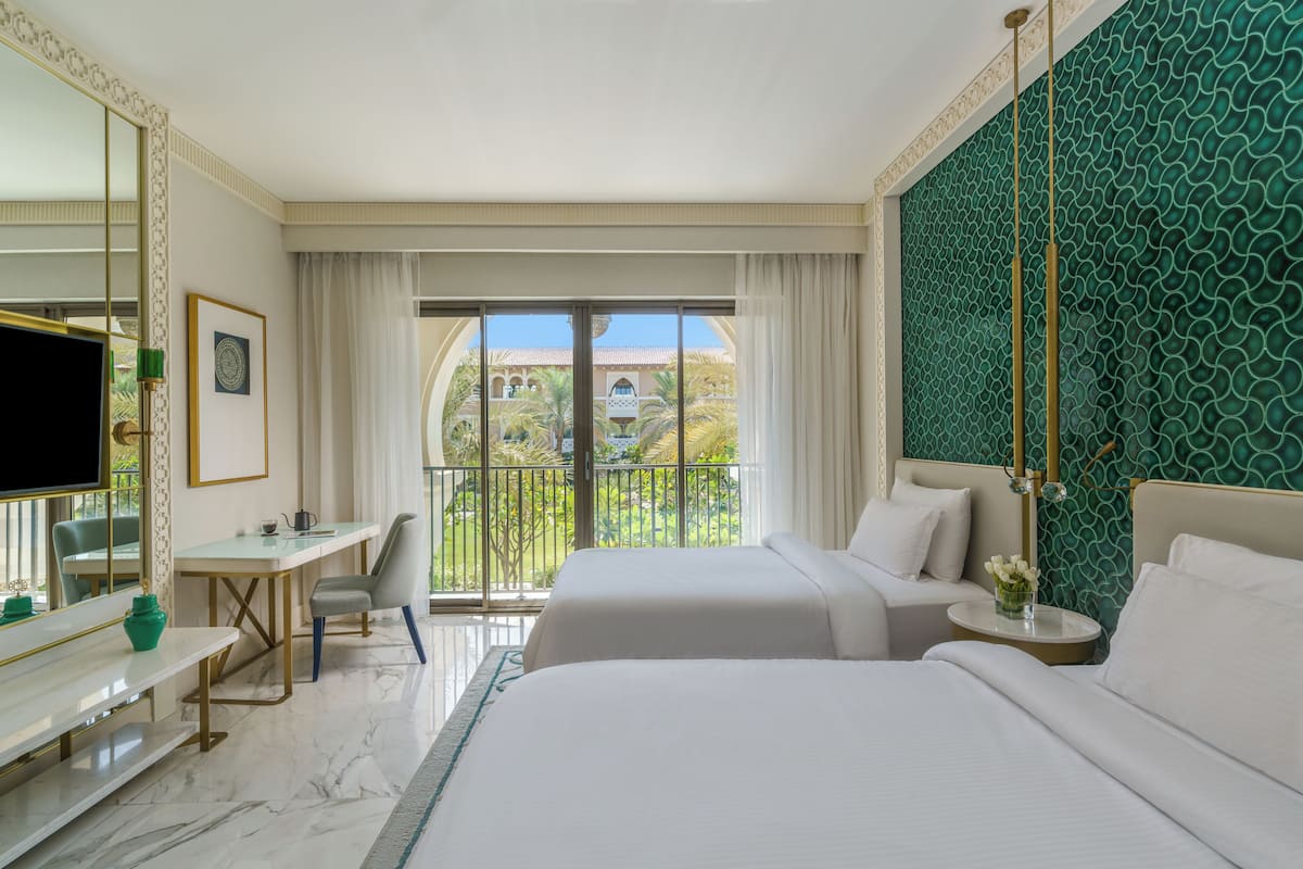 Une chambre d’hôtel avec deux lits, un bureau, une chaise, une télévision et un balcon offrant une vue sur des palmiers et un immeuble.