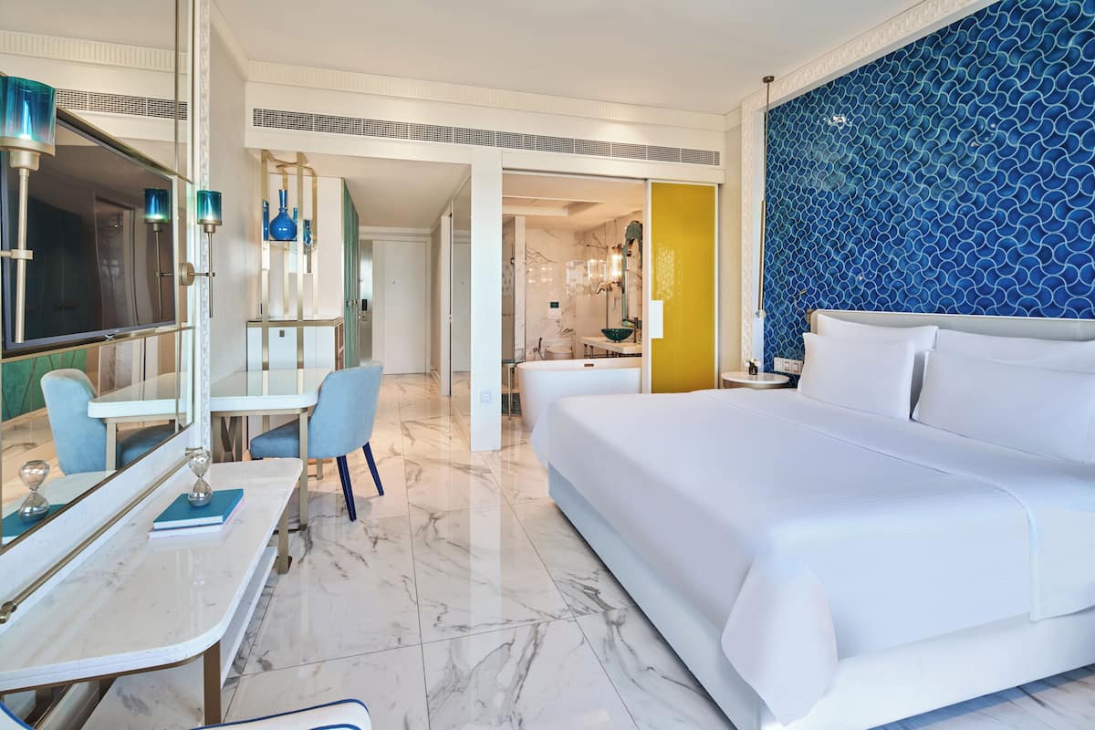 Une chambre d’hôtel moderne dotée d’un grand lit, d’un bureau avec une chaise, d’un miroir et d’une salle de bain avec une baignoire et une douche.