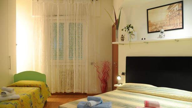 Habitación de hotel con dos camas, una silla verde, una ventana con cortinas translúcidas, un estante montado en la pared con objetos decorativos, y un cuadro enmarcado sobre el estante.