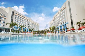 Ett poolområde vid hotellet med solstolar och parasoller, omgivet av palmer och moderna byggnader.