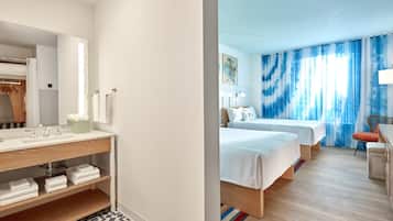 A hotel room with a wooden vanity, a large mirror, two beds, a chair, and a window with blue curtains.