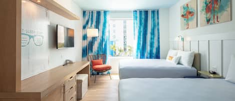 Habitación de hotel con dos camas, un escritorio, una silla, un televisor y obras de arte en las paredes.