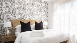 Un lit bien fait, avec du linge de lit blanc et deux oreillers noirs, une tête de lit en bois, une table de chevet avec une lampe et un papier peint à motifs.