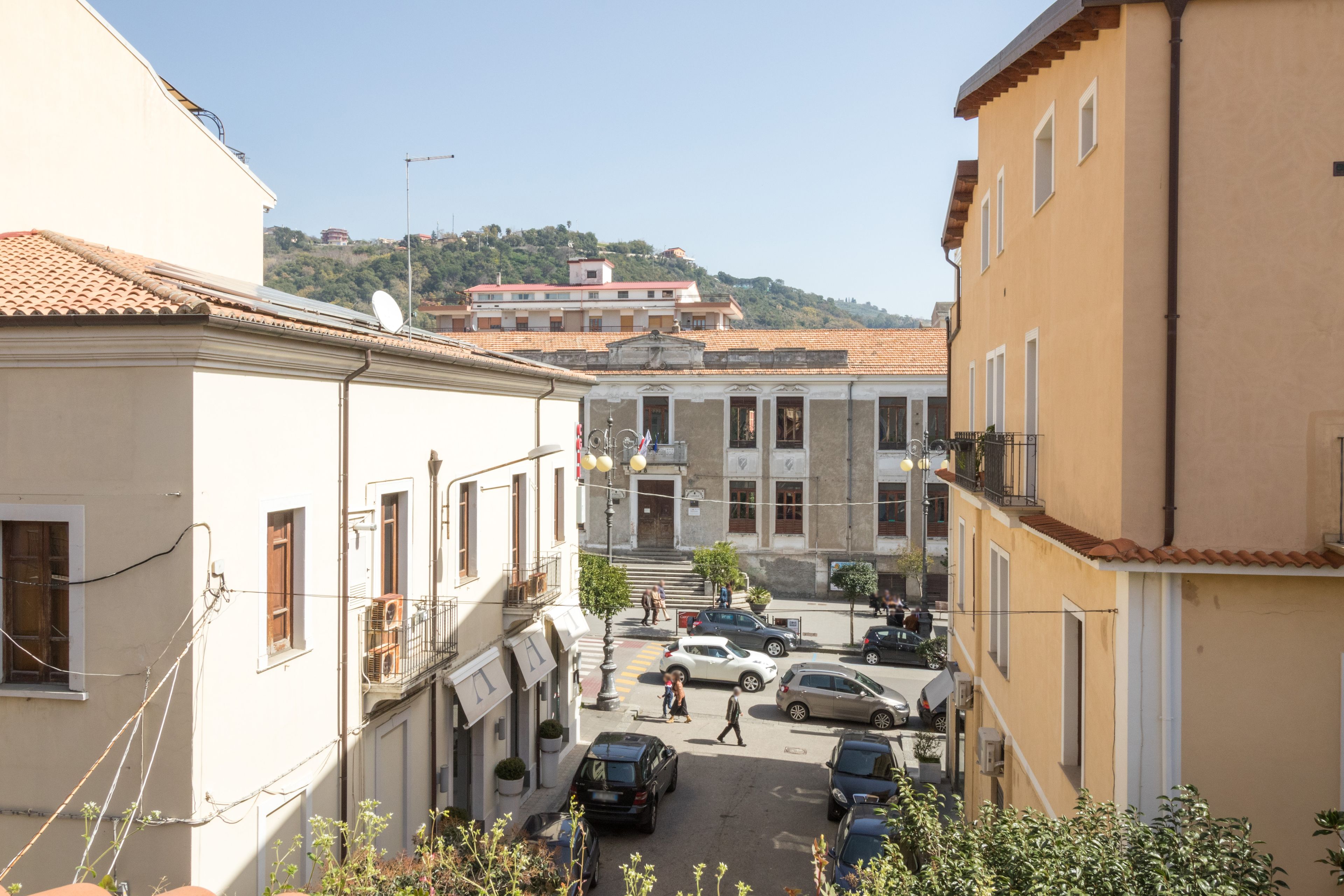 A narrow street with buildings on either side, a few parked cars, and pedestrians.