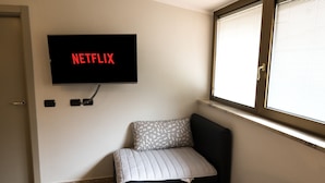 Quarto com cama, televisão fixada na parede exibindo o logotipo da Netflix, janela com persianas e porta.