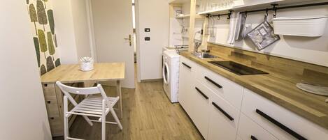 Cocina compacta con encimera de madera, armarios blancos, lavadora y un pequeño comedor con una silla.