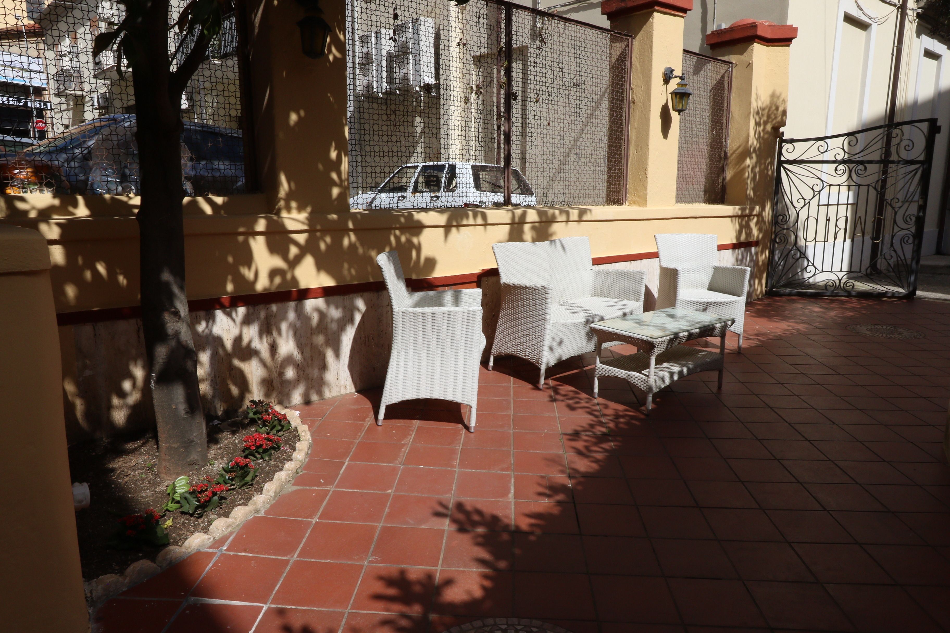 Eine Terrasse im Freien mit weißen Korbmöbeln, gefliestem Boden und Blick auf eine Straße mit einem Auto.