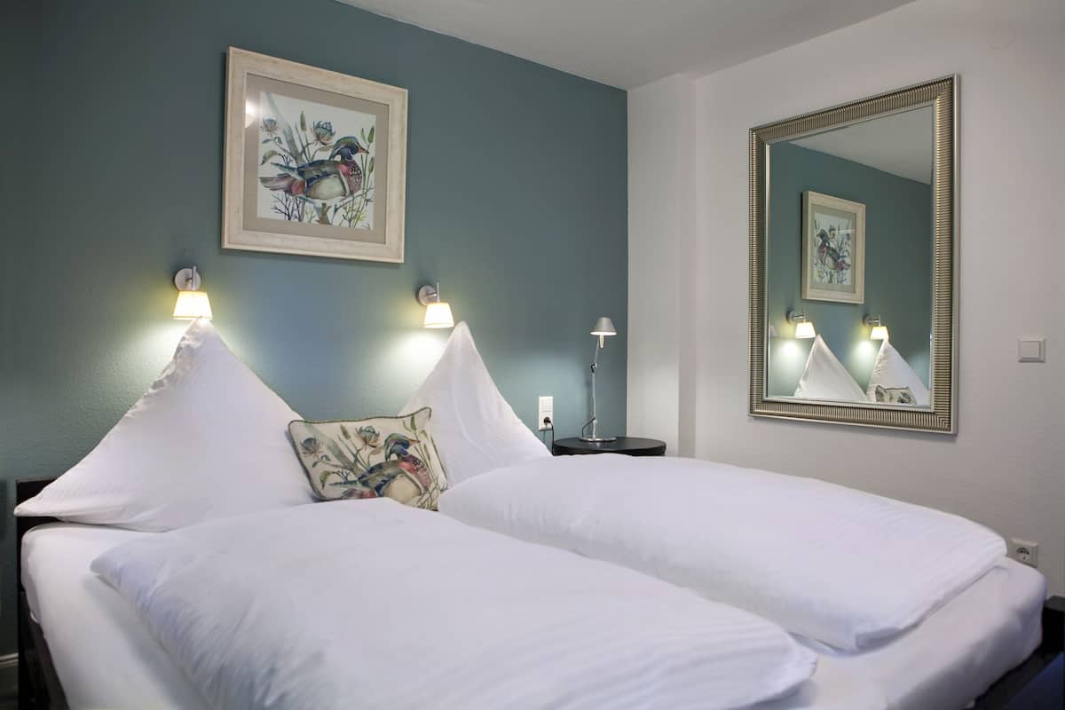 Una cama bien hecha con ropa de cama blanca, una obra de arte floral enmarcada encima y un espejo grande en la pared.