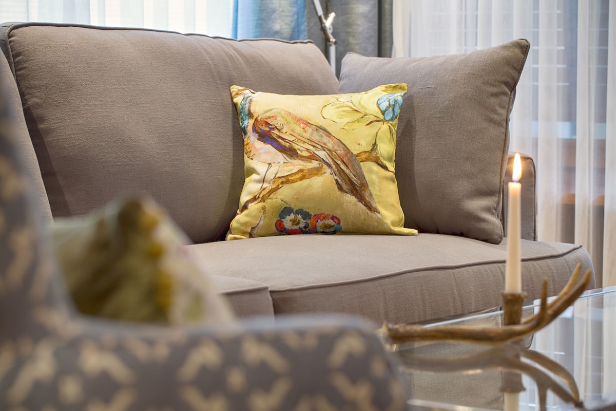 Una sala moderna con sofá, un cojín decorativo, una mesa de vidrio con vela y una ventana con persianas.