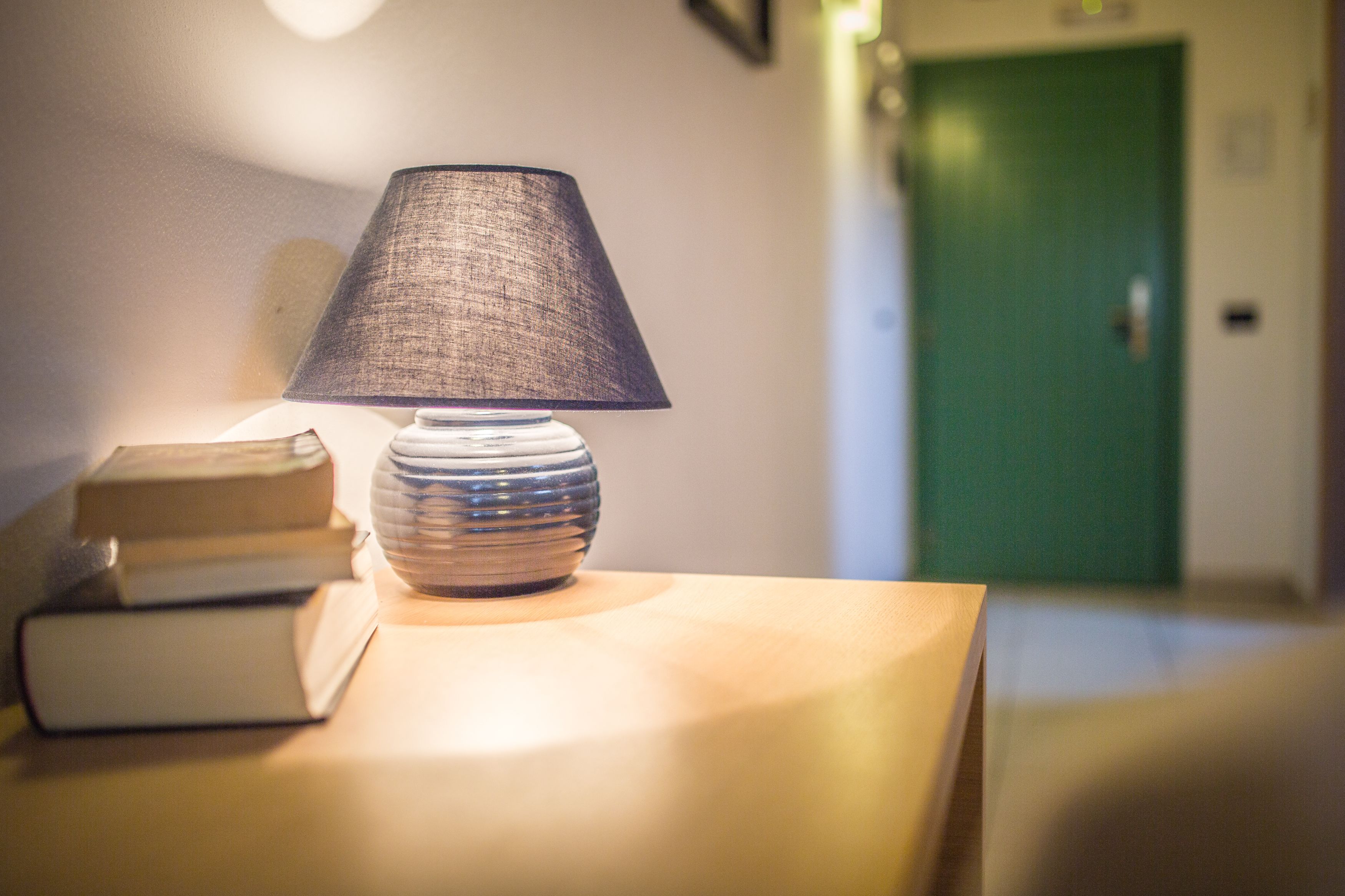 A lamp on a bedside table with books beside it, a green door in the background, and a hallway leading to it.