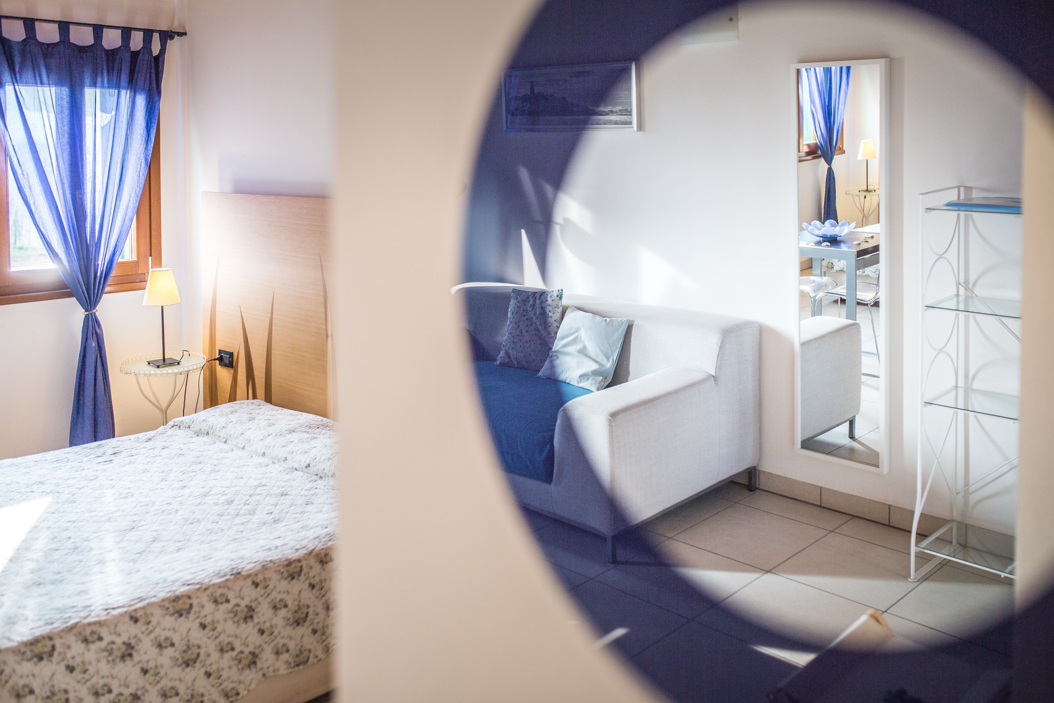 A bedroom with a bed, a bedside table, a lamp, a window with blue curtains, and a view of a cityscape through the window.