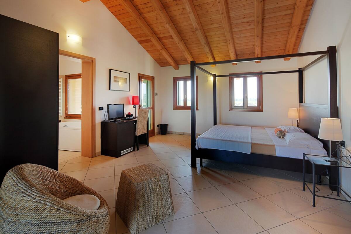 Une chambre spacieuse avec un lit à baldaquin, un bureau avec un ordinateur et un fauteuil en osier.