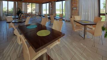 Un vaste espace extérieur aménagé pour les repas, avec plusieurs tables et chaises, de grandes fenêtres et une vue sur le paysage environnant.