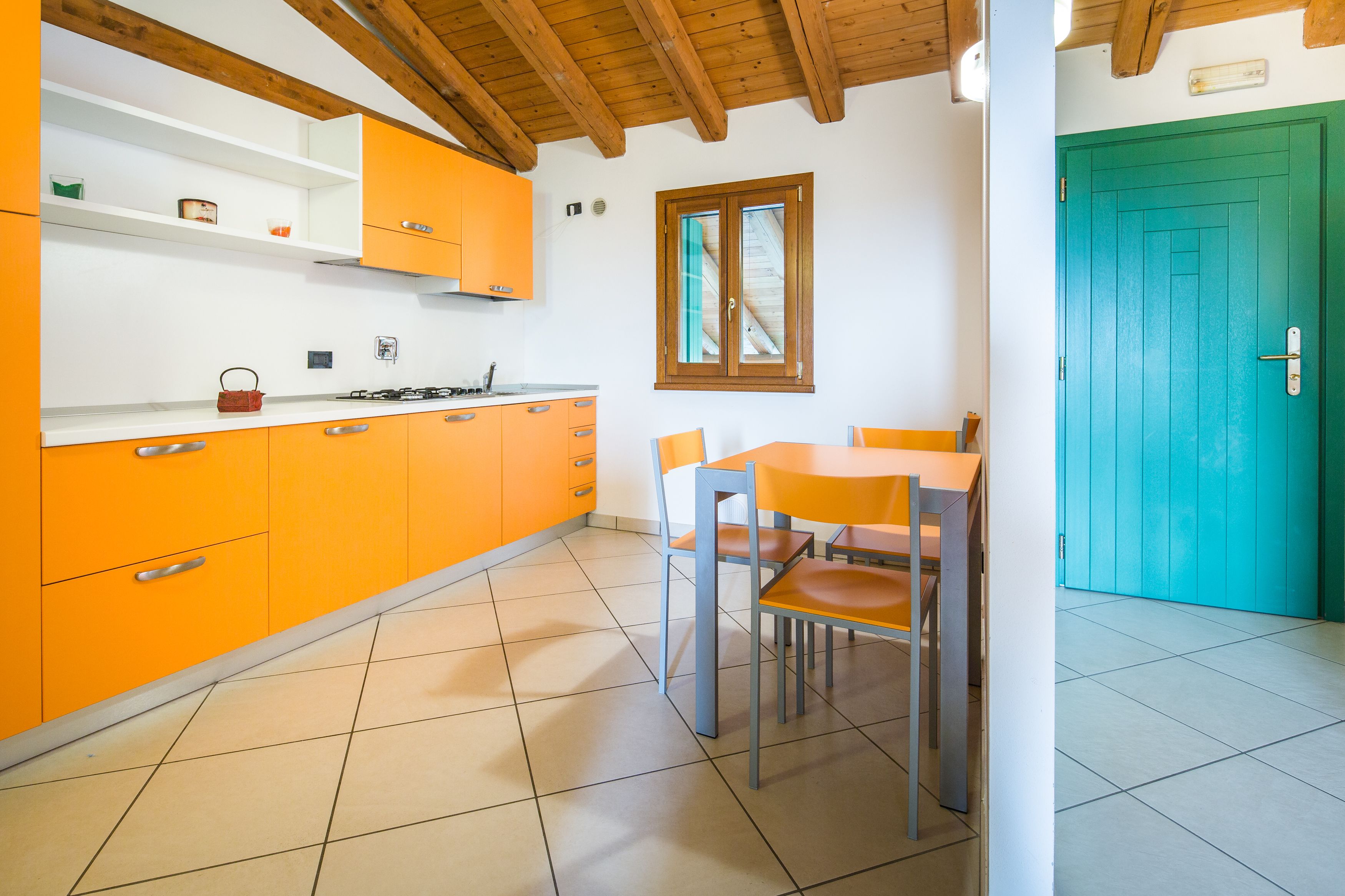Une cuisine avec des meubles de couleur orange, un plafond en bois, une table à manger avec des chaises et une porte verte.