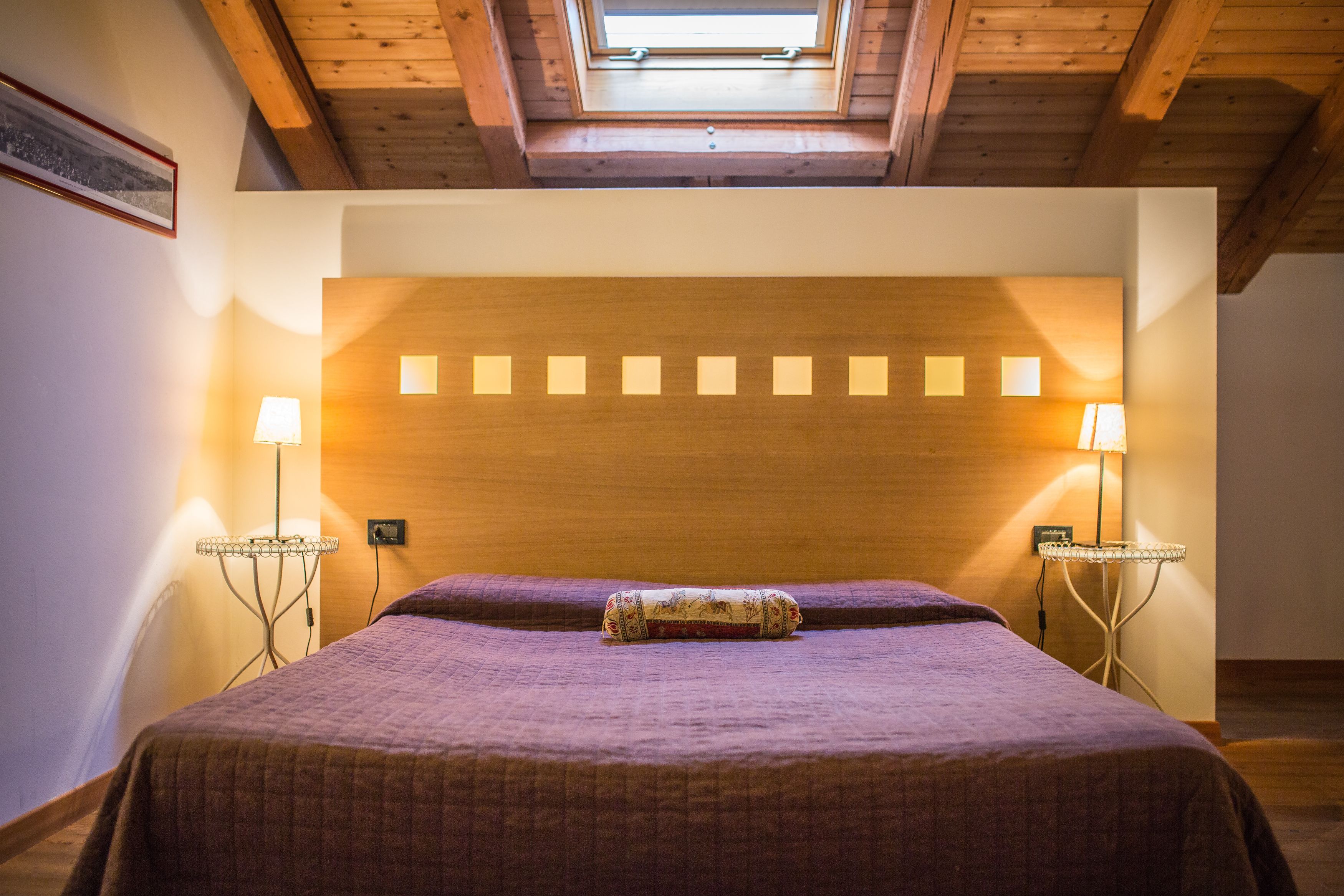 A bedroom with a wooden headboard, a bed with a purple bedspread, two bedside lamps, and a skylight above.