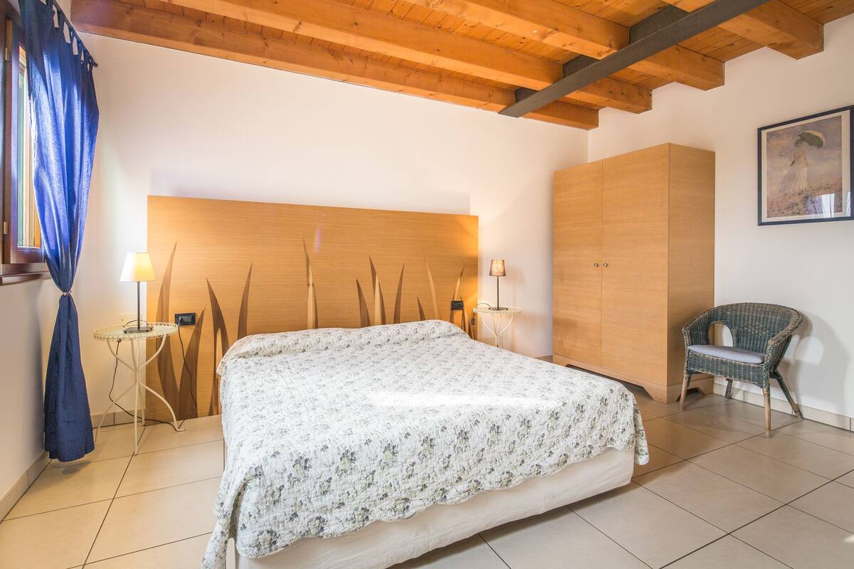 Une chambre avec un grand lit, une tête de lit en bois, deux lampes de chevet, un fauteuil en osier et un tableau accroché au mur.