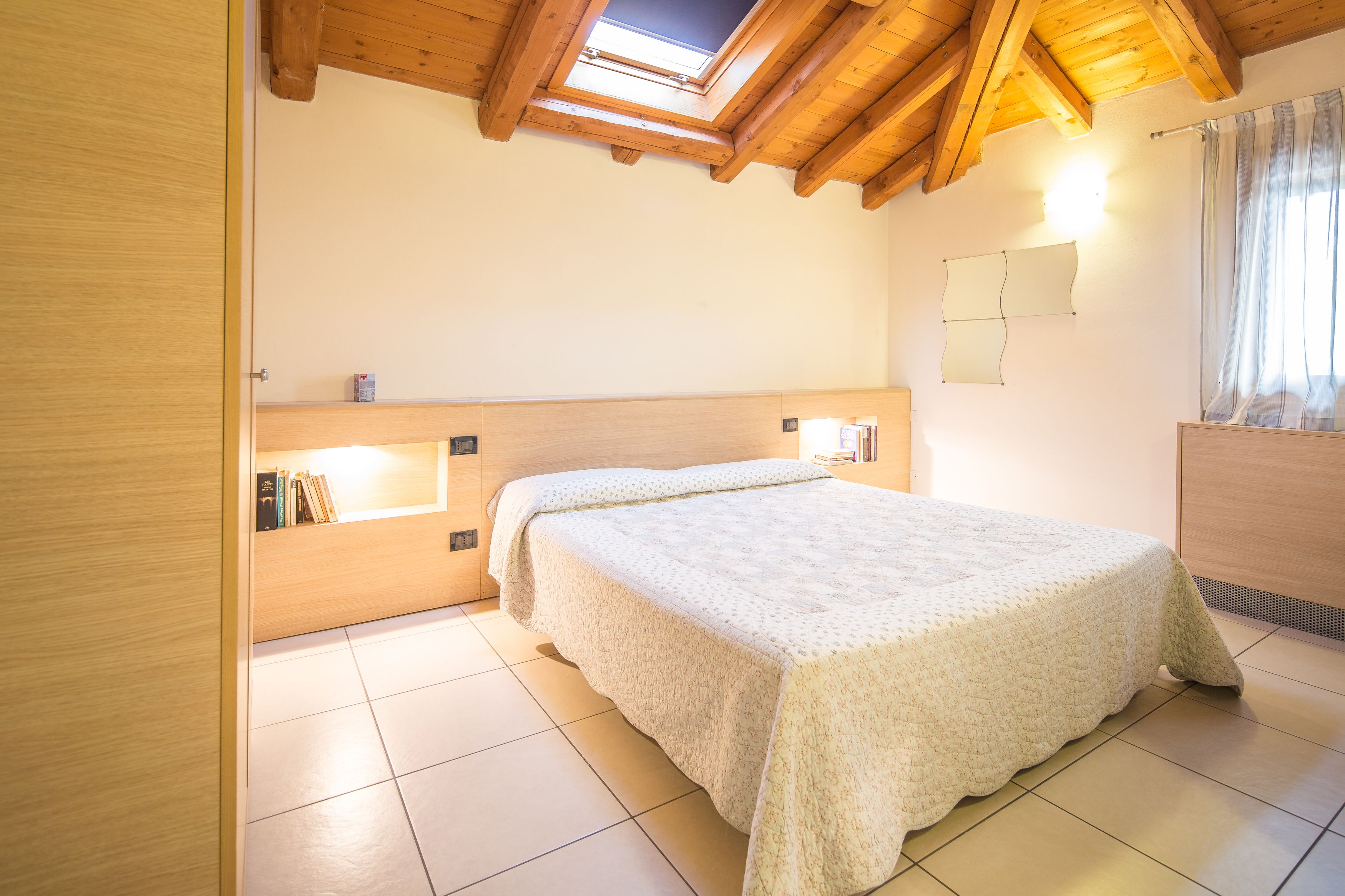Ein Schlafzimmer mit Holzbalkendecke, einem Oberlicht, einem Bett mit weißer Bettdecke, einem Nachttisch mit Büchern und einer wandmontierten Leuchte.