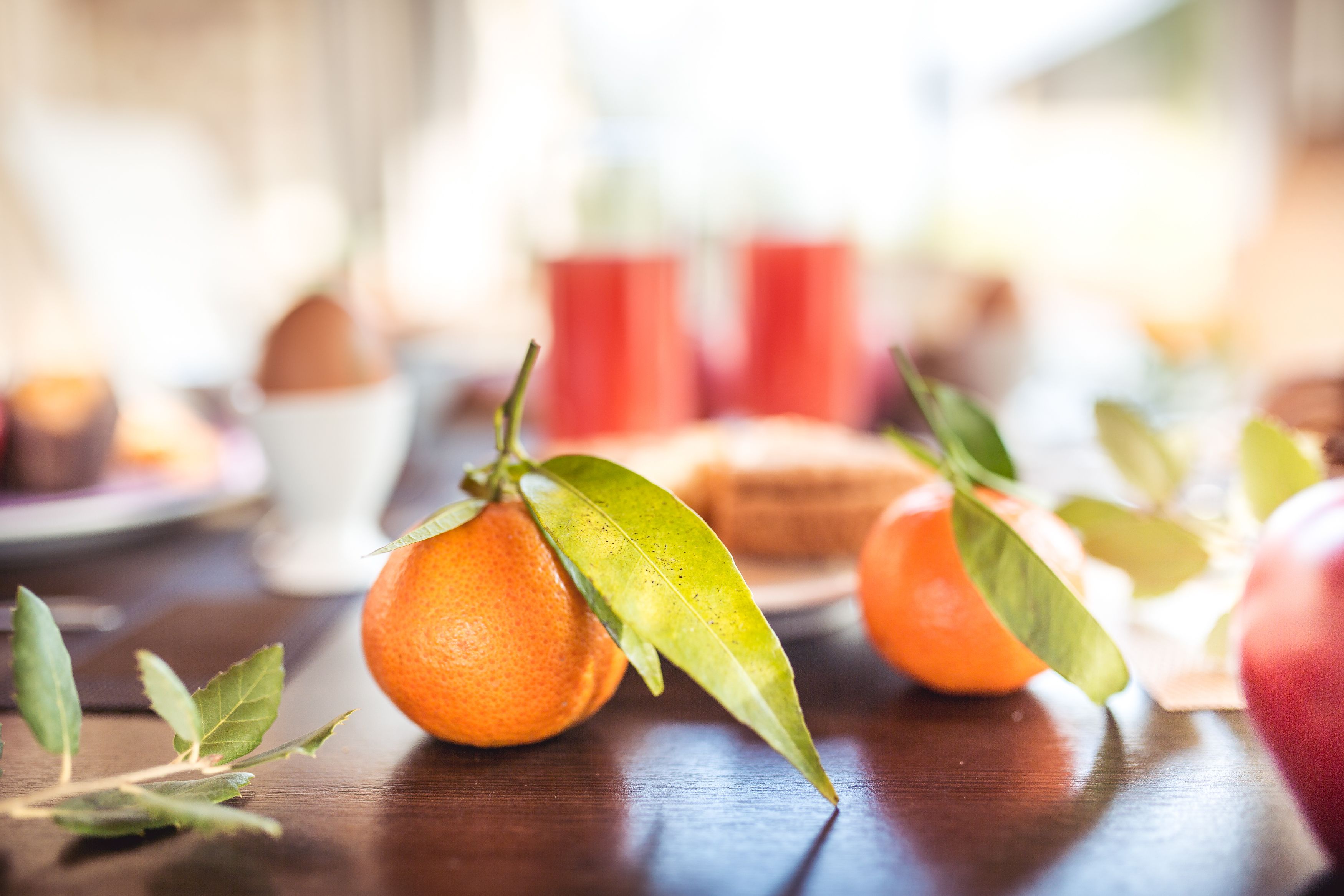 Una mesa de madera con naranjas y hojas verdes, una taza blanca, y tazas rojas al fondo.
