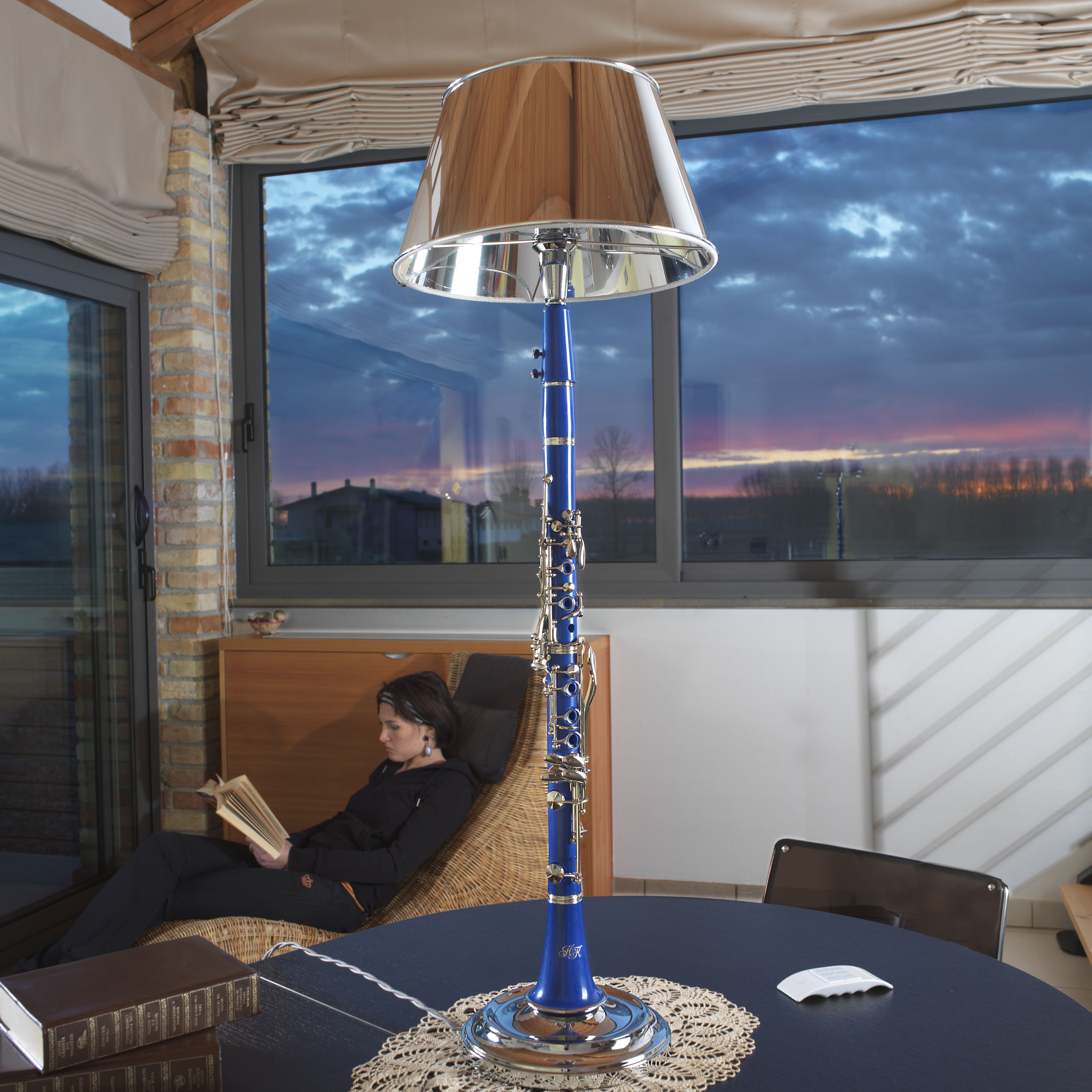 Una habitación con un ventanal grande, una lámpara de mesa azul, una silla de mimbre y una persona leyendo un libro.