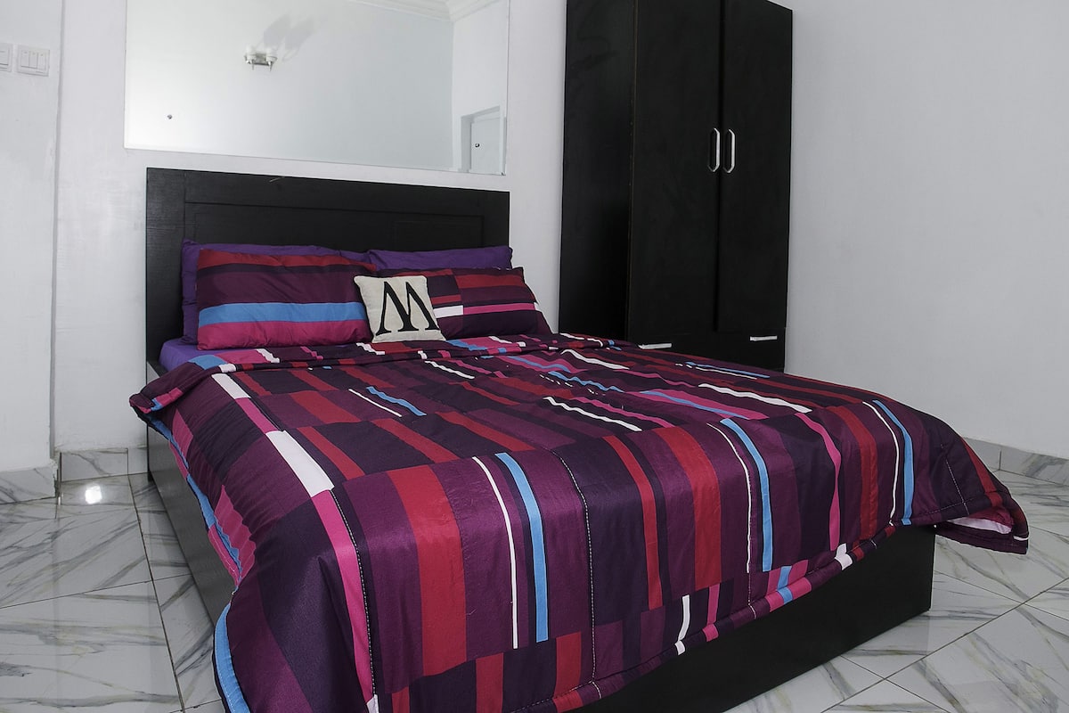 A bed with a striped bedspread, a pillow with a large 'M', and a black wardrobe in a room with white walls and a tiled floor.