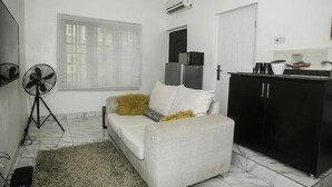 A compact living space with a sofa, a small kitchen area, a standing fan, and a window with blinds. - Capital & County Apartment Chevron (Lekki)