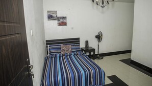 A bedroom with a bed, a nightstand, a wall-mounted fan, and a door. - Capital & County Apartment Chevron (Lekki)
