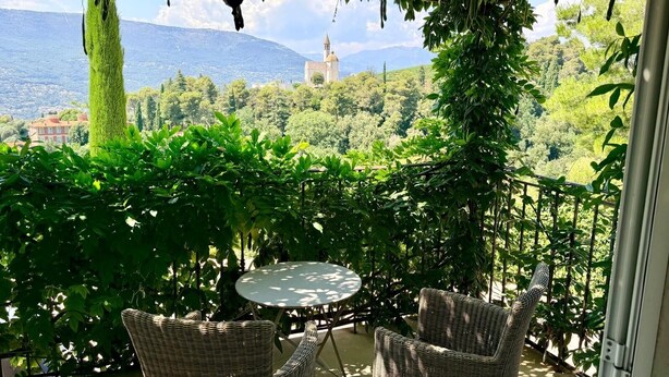 A balcony with wicker furniture and a table, overlooking a lush garden and a distant church. - Villa Kilauea B&B (Nice)