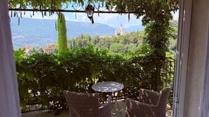 A balcony with wicker furniture, a round table, and a view of a mountainous landscape with a church. - Villa Kilauea B&B (Nice)