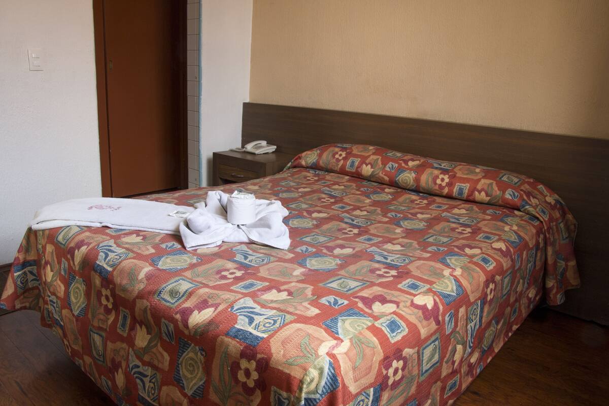 Un letto rifatto con cura, una coperta a fantasia, un comodino con un telefono e una porta sullo sfondo.
