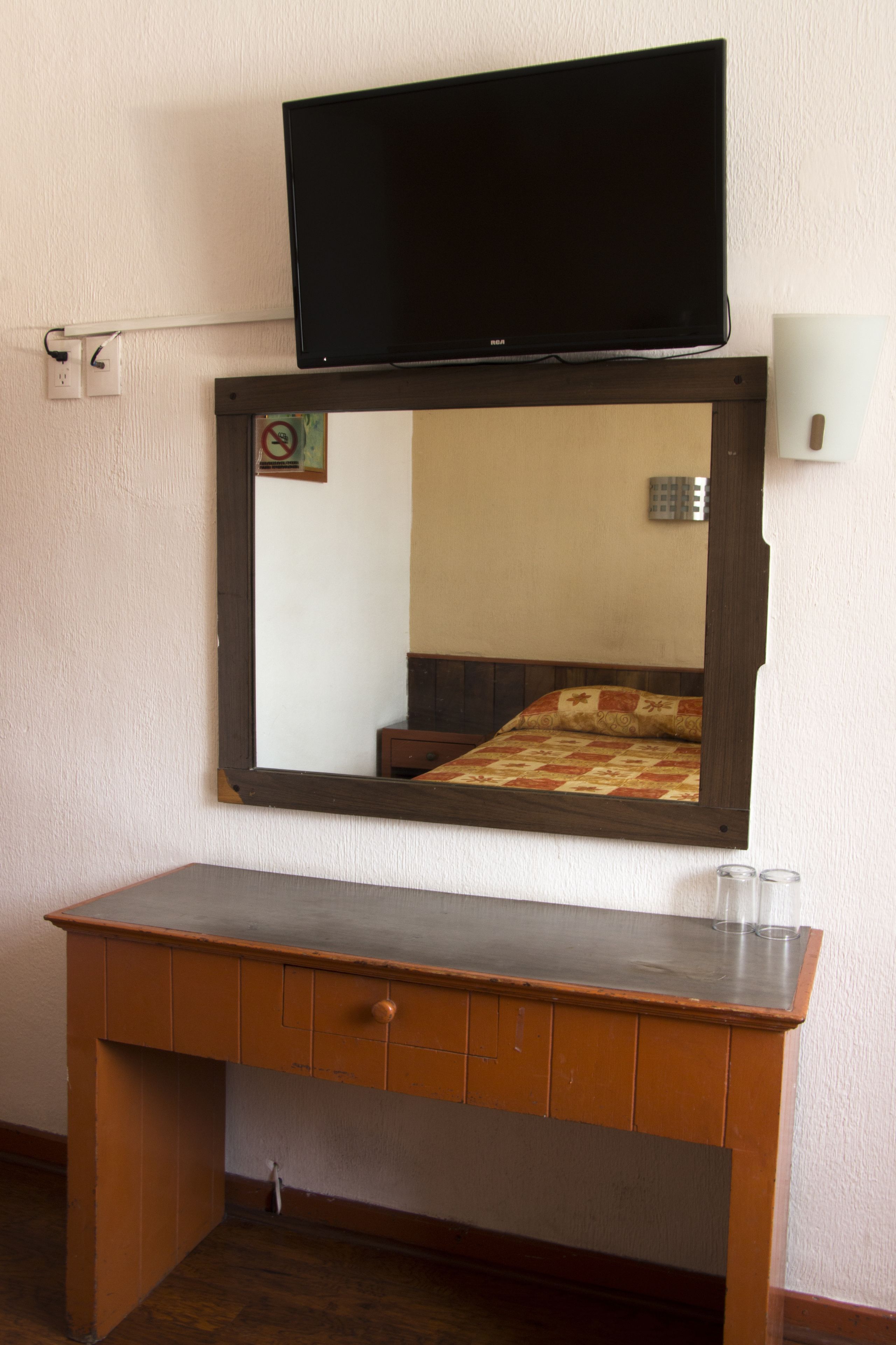 A room with a bed, a wooden desk, a flat-screen TV mounted above the desk, and a wall socket.