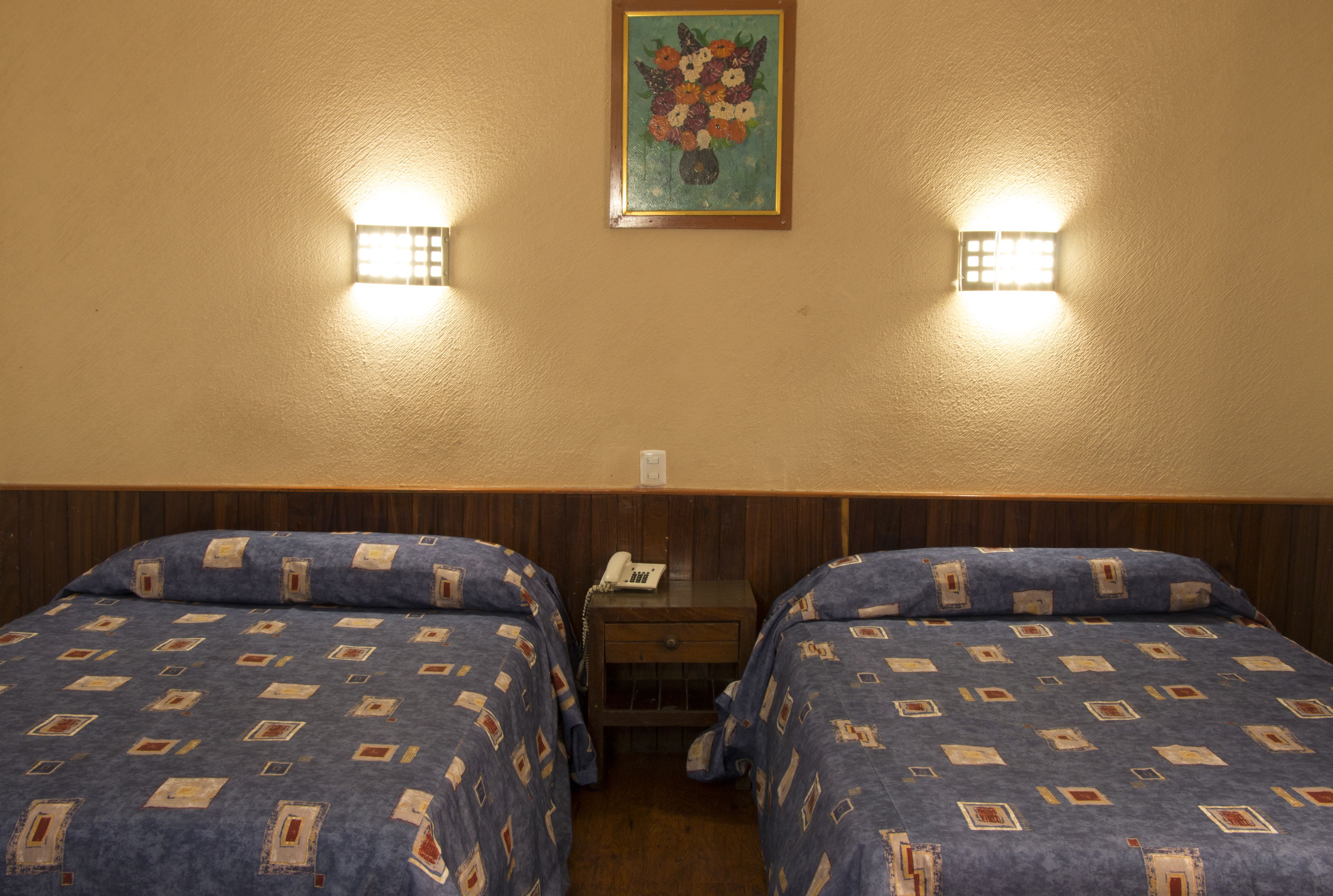 A room with two beds, a nightstand with a phone, and a wall-mounted light fixture.