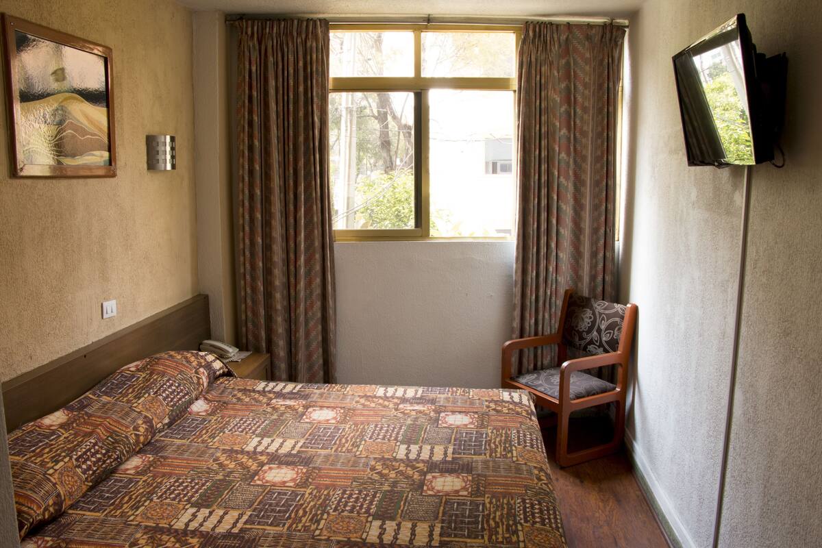 Una stanza con un letto, una sedia, una finestra con tende, una TV a parete e un quadro.