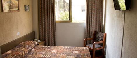 Un dormitorio con cama, una silla, una ventana con cortinas, un cuadro en la pared y un televisor montado en la pared.