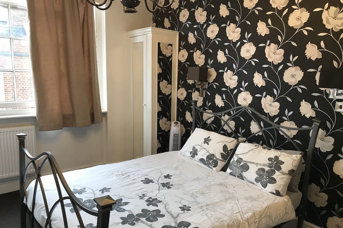 Un dormitorio con cama, radiador, ventana con cortinas y pared con papel tapiz floral.