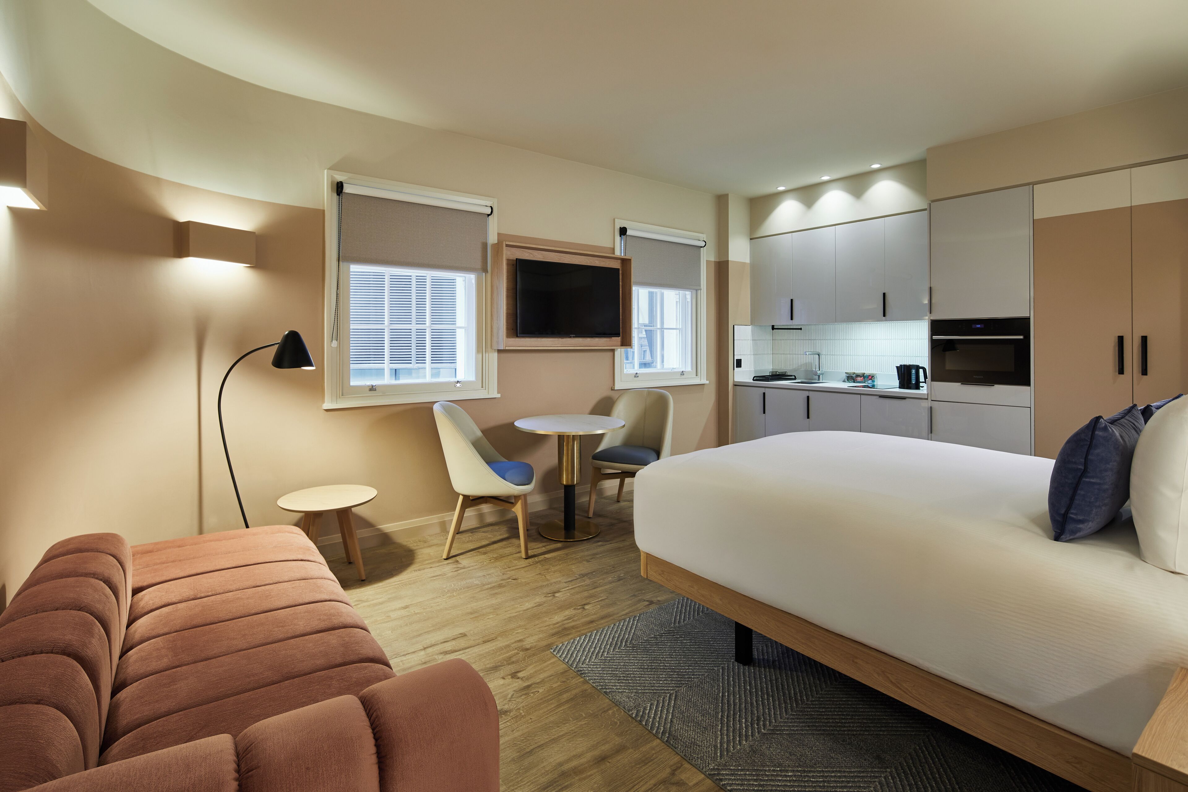 A modern hotel room with a large bed, a sofa, a dining table, and a kitchenette.