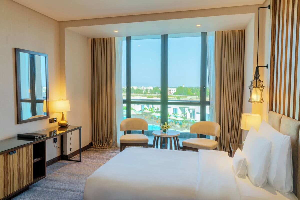 Habitación de hotel con una cama grande, dos sillones, una mesita con lámpara, un escritorio con lámpara, y un ventanal con vistas a edificios y vegetación.