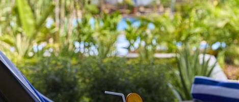 A glass table with a banana and a drink, a towel, and a striped chair with a view of a pool and greenery.