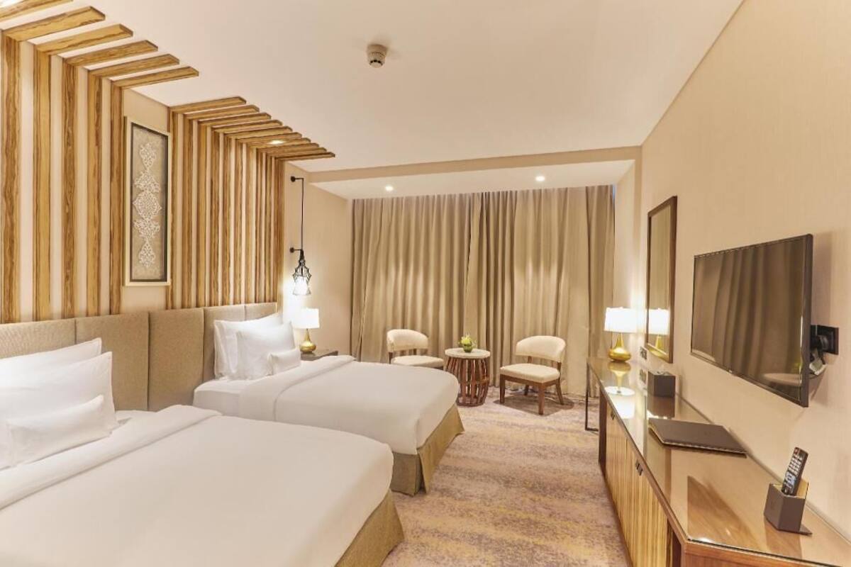 Habitación de hotel con dos camas, un escritorio con televisor, un espejo y una zona de estar con sillas y una mesita.