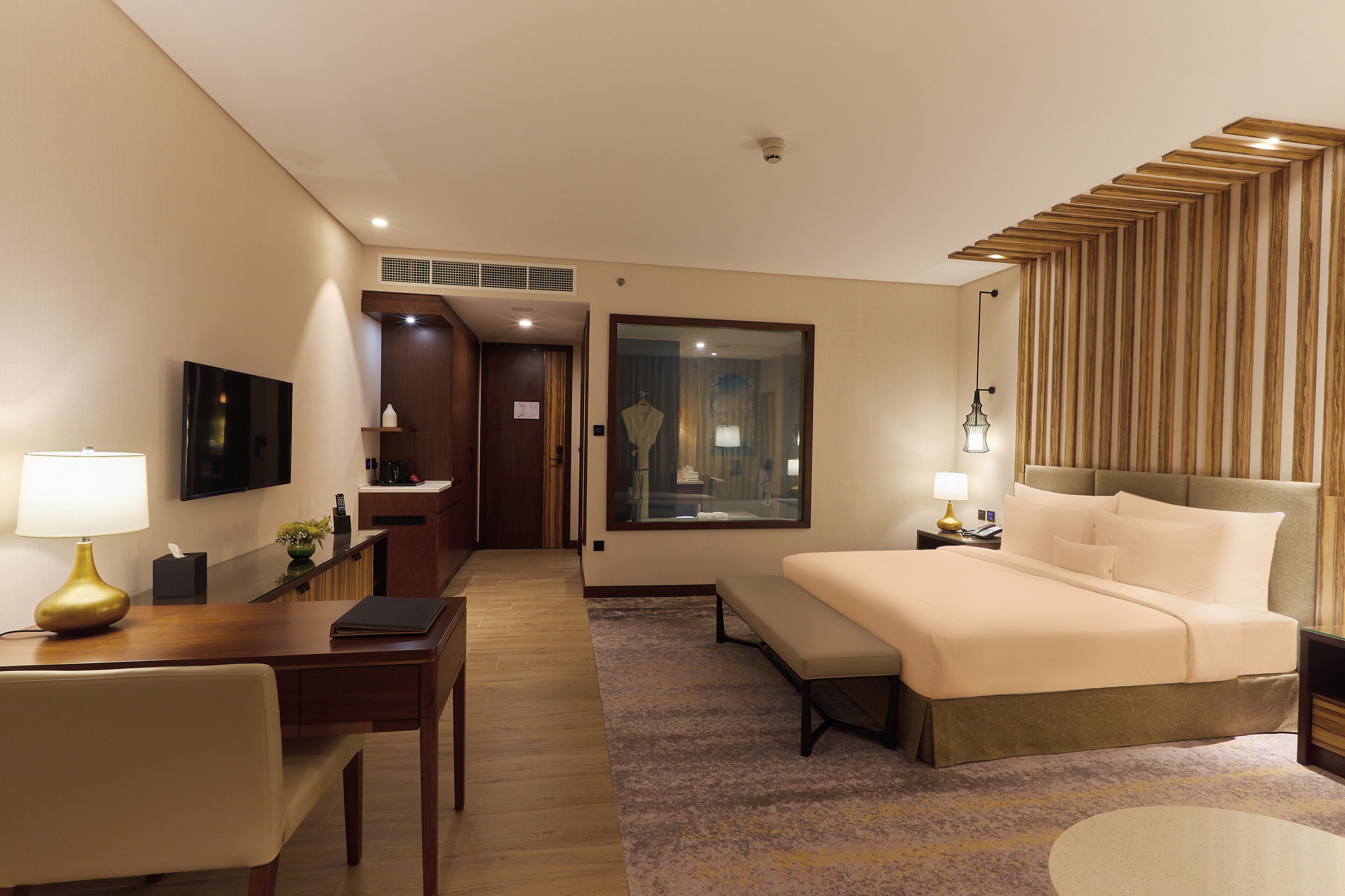 Quarto de hotel com cama, mesa de cabeceira, luminária, escrivaninha, cadeira e vista para o corredor.