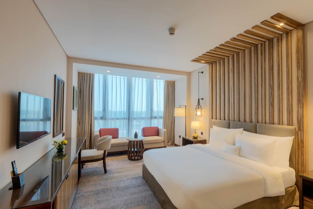 Habitación de hotel con una cama grande, televisión, zona de estar con mesa y vistas al exterior a través de amplias ventanas.