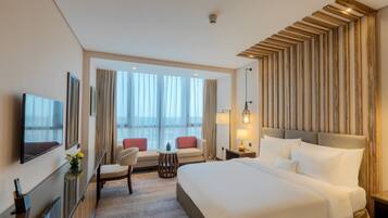 Habitación de hotel con una cama grande, televisión, zona de estar con mesa y vistas al exterior a través de amplias ventanas.