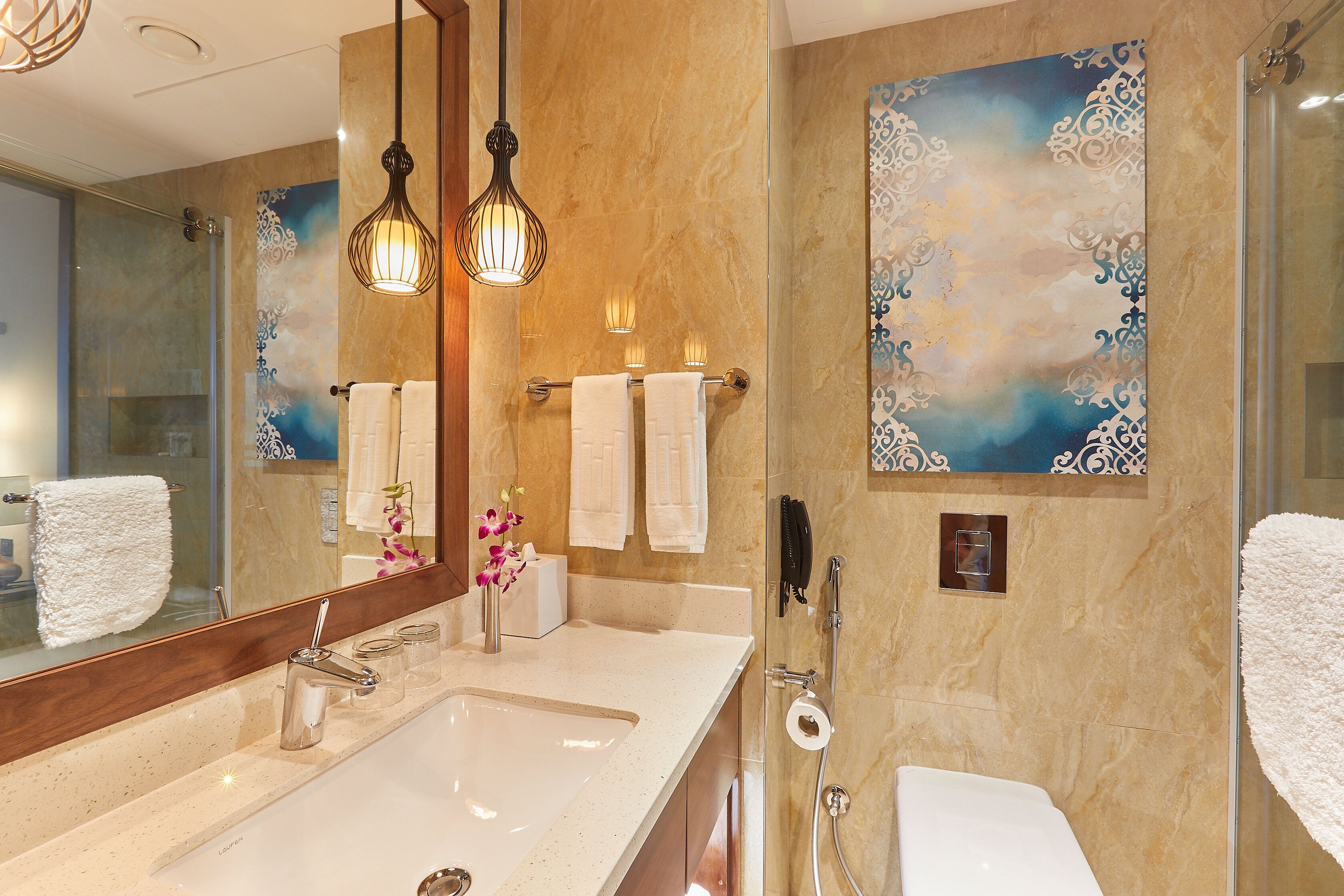 Un baño moderno con un espejo grande, un lavamanos, cabina de ducha con mampara de vidrio y detalles decorativos en la pared.