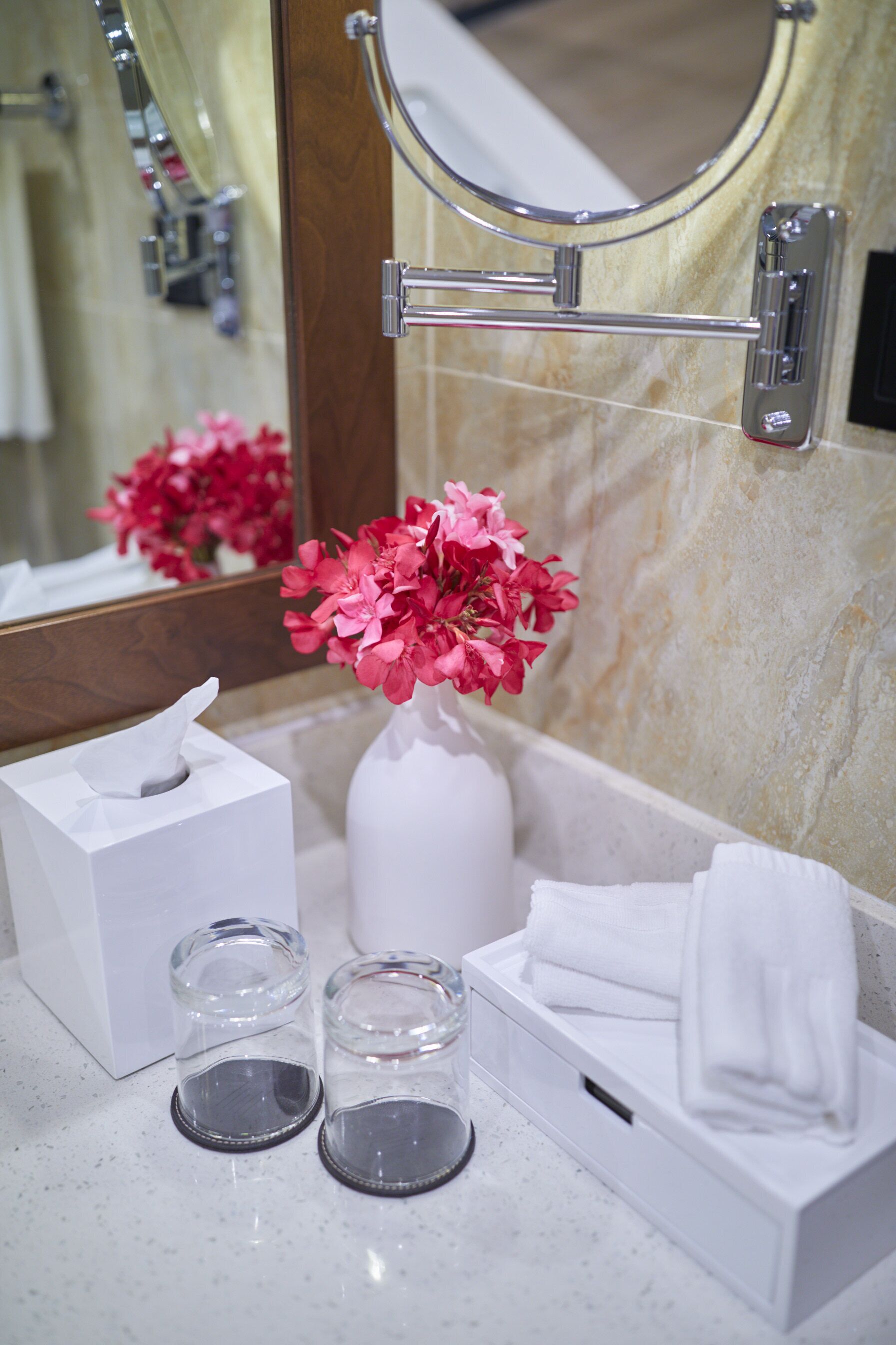 A bathroom with a marble sink, a mirror, a tissue box, a vase with pink flowers, and a towel rack.