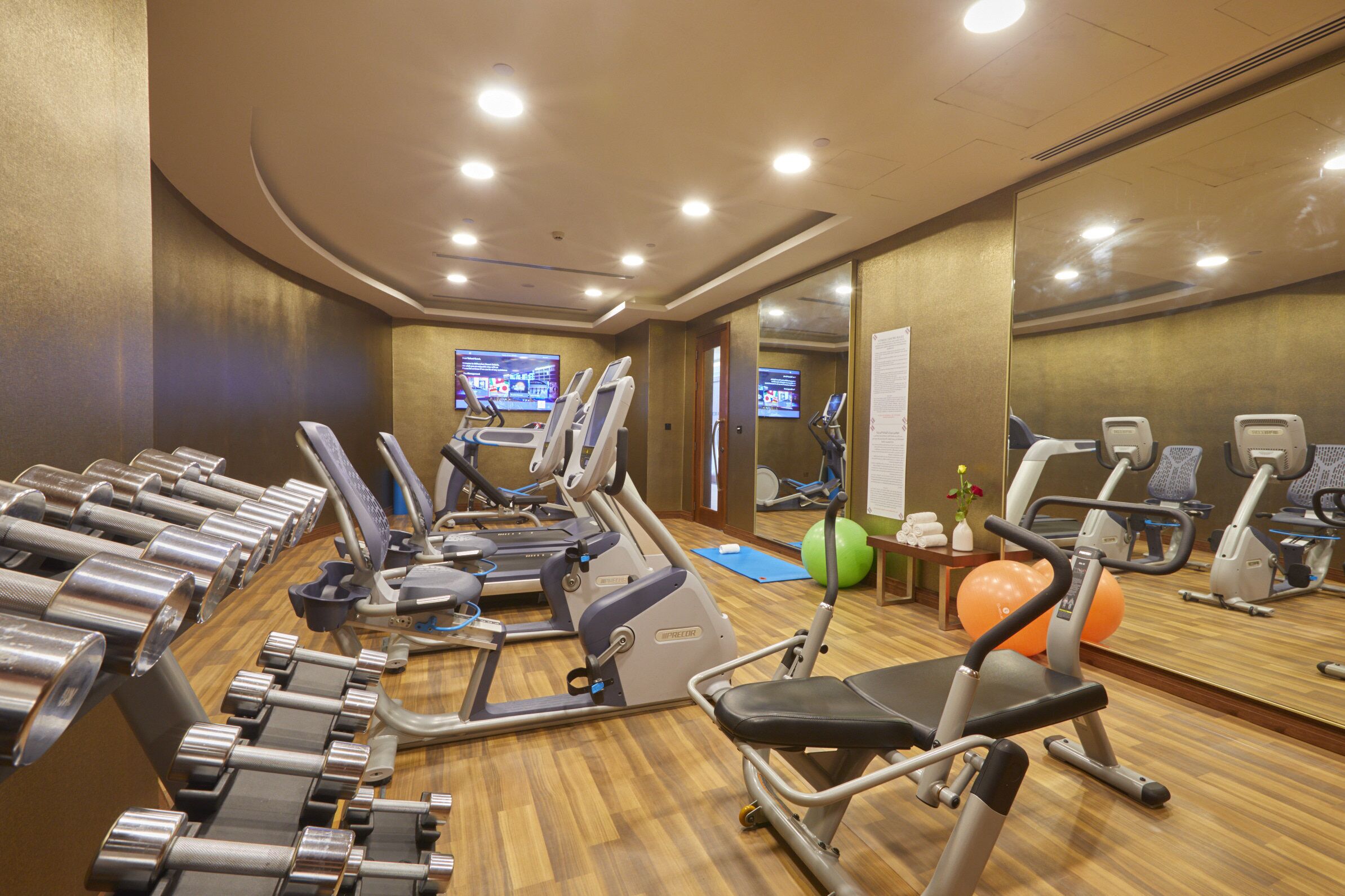 Un gimnasio con varias máquinas de ejercicio, suelo de madera y pared de espejos.