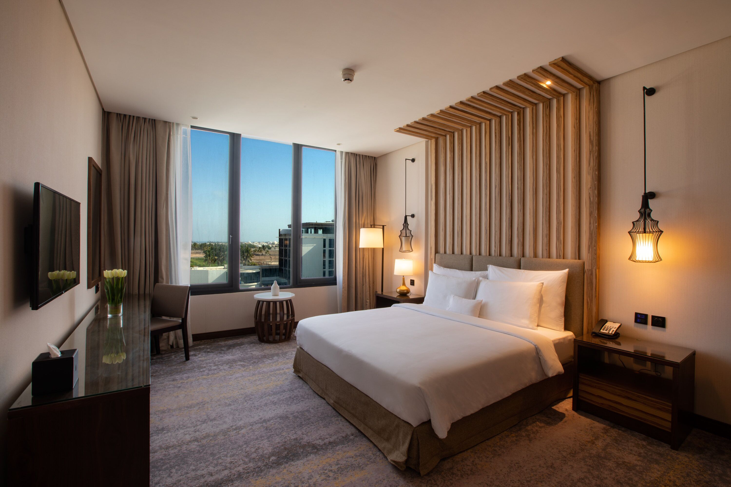 A hotel room with a large bed, a desk with a lamp, a chair, a small table, and a view of the city through large windows.