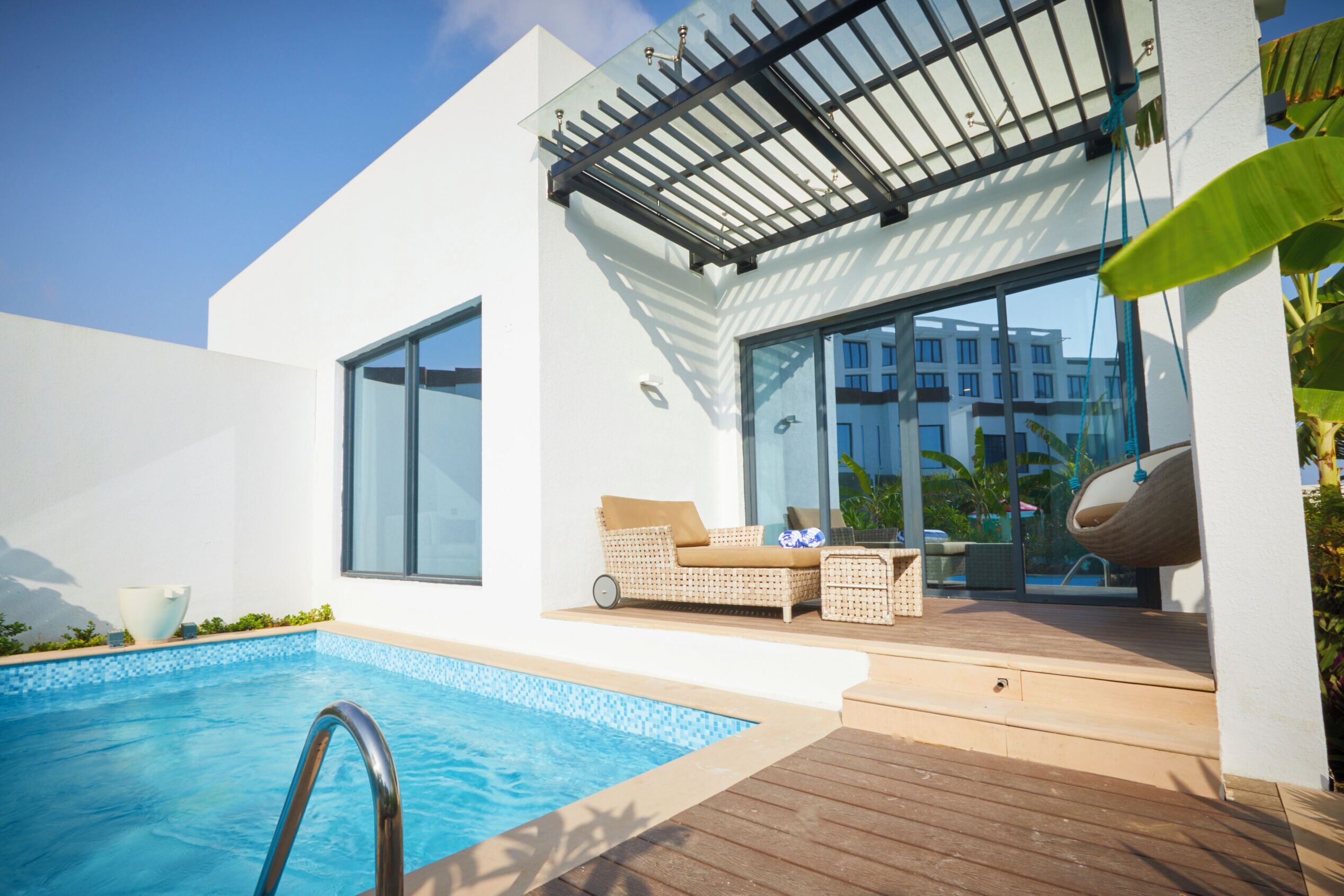 Un espace piscine extérieur moderne, doté d'une balustrade en verre, de mobilier en osier et offrant une vue sur un bâtiment en arrière-plan.