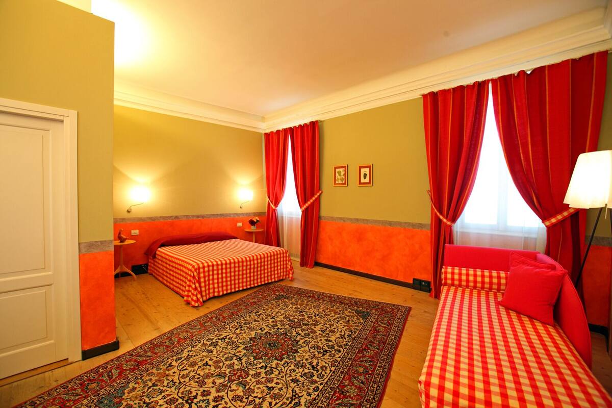 A hotel room with two beds, red curtains, a patterned rug, and a small table with a lamp.