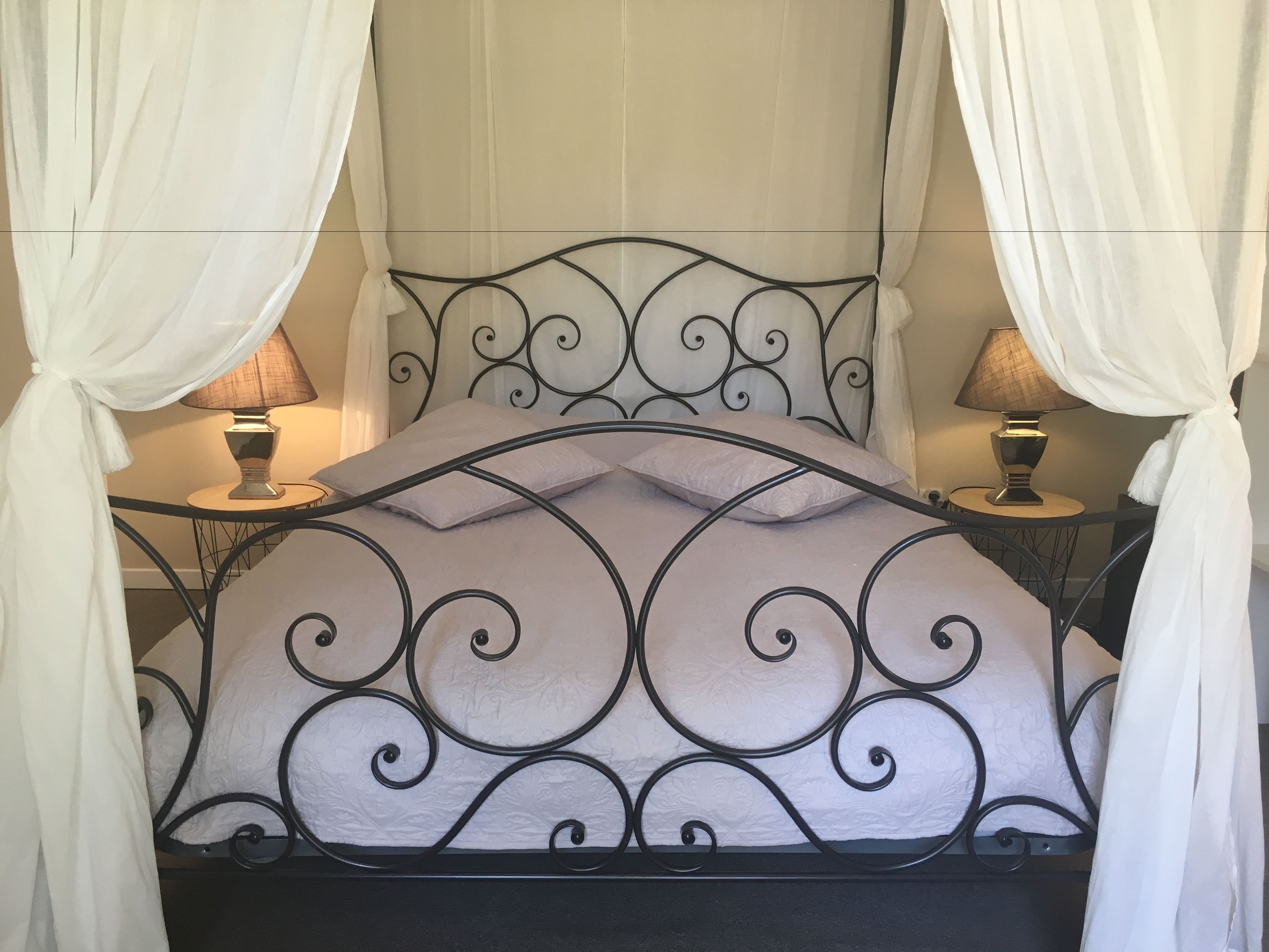 A bed with a black metal frame and white bedding, flanked by two bedside lamps and draped with white curtains.